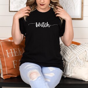 May include: A woman wearing a black t-shirt with the word "Witch" in white script. She is sitting on a bench with pillows behind her.