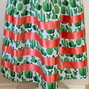 May include: A white skirt with a green cactus print and a coral ribbon trim. The skirt is gathered at the waist and has a flowy silhouette.