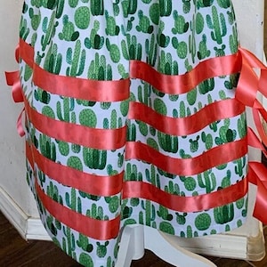 May include: A white skirt with a green cactus print and coral ribbon trim. The skirt has an elastic waistband and is a midi length.