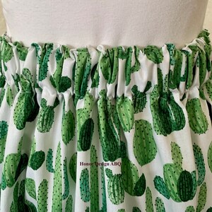 May include: White fabric skirt with a green cactus print. The skirt has an elastic waistband and is gathered at the top.