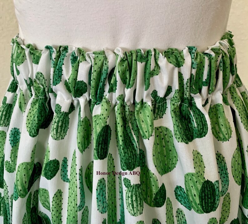 May include: White fabric skirt with a green cactus print. The skirt has an elastic waistband and is gathered at the top.