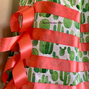 May include: A white fabric skirt with a green cactus print and coral ribbon trim. The skirt has a gathered waist and is decorated with multiple coral ribbons tied in bows.