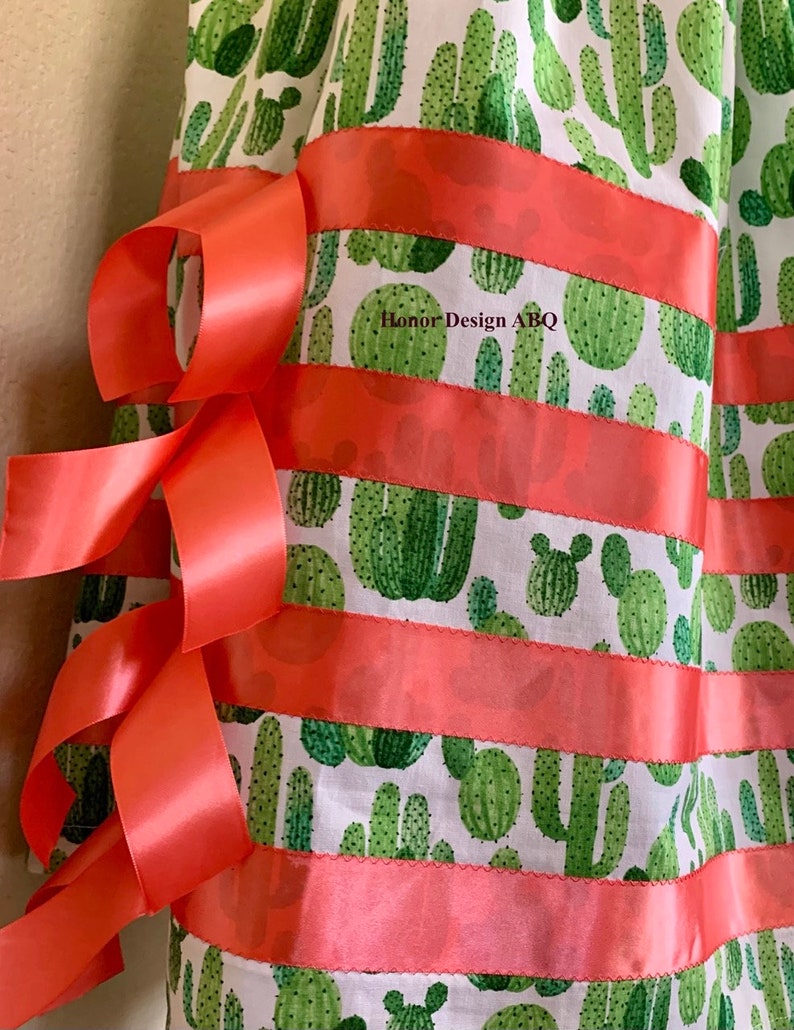 May include: A white fabric skirt with a green cactus print and coral ribbon trim. The skirt has a gathered waist and is decorated with multiple coral ribbons tied in bows.