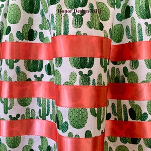 May include: A white fabric with a green cactus print and coral colored ribbon trim. The fabric has a repeating pattern of different sized cacti.