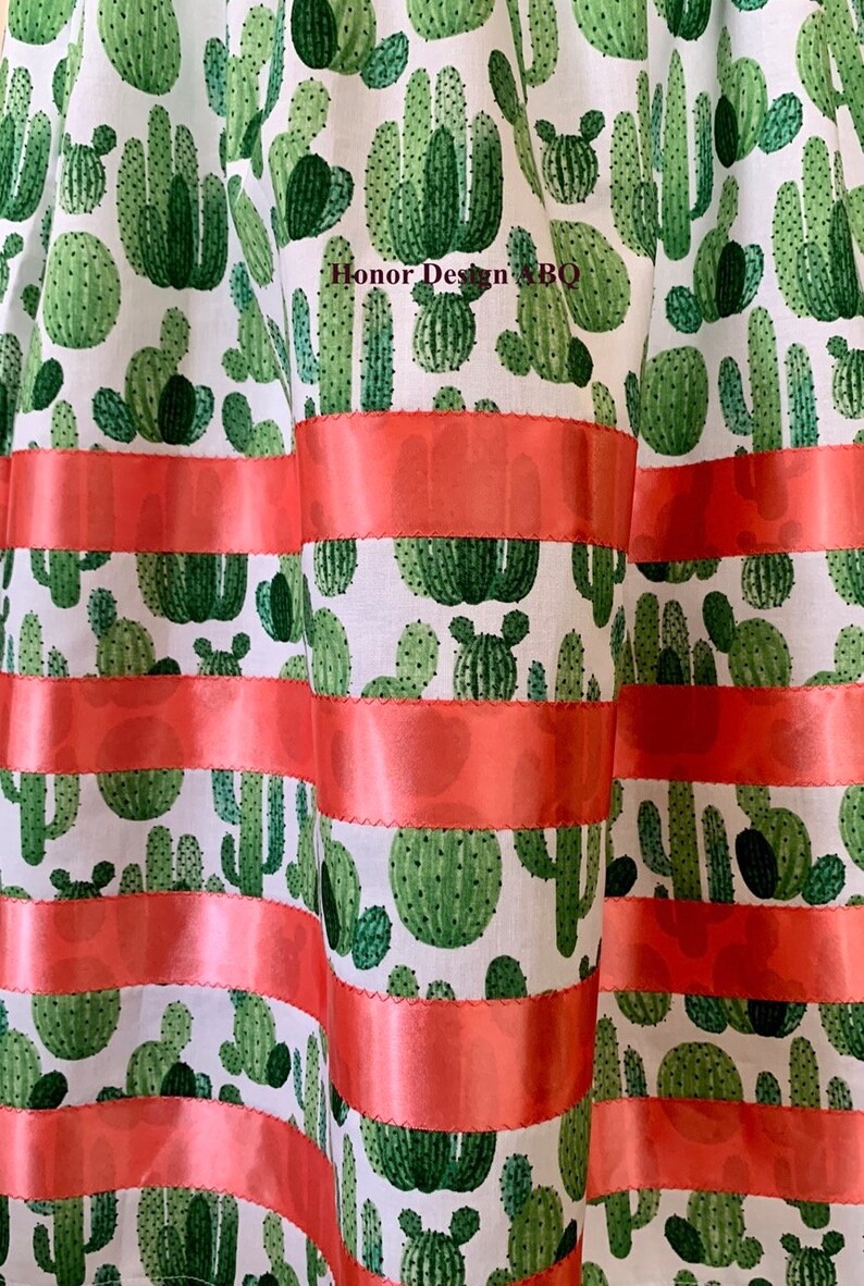 May include: A white fabric with a green cactus print and coral colored ribbon trim. The fabric has a repeating pattern of different sized cacti.