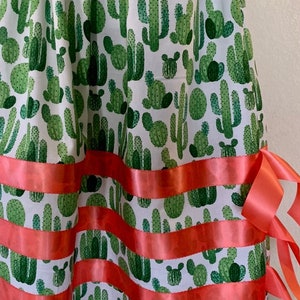 May include: A white skirt with a green cactus print and red ribbon trim. The skirt has a gathered waist and a full skirt. The skirt is made of cotton and is perfect for a summer party or a casual day out. Honor Design ABQ is printed on the skirt.