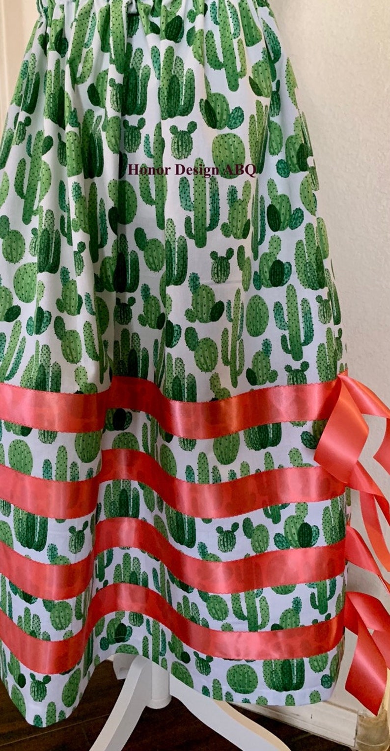 May include: A white skirt with a green cactus print and red ribbon trim. The skirt has a gathered waist and a full skirt. The skirt is made of cotton and is perfect for a summer party or a casual day out. Honor Design ABQ is printed on the skirt.
