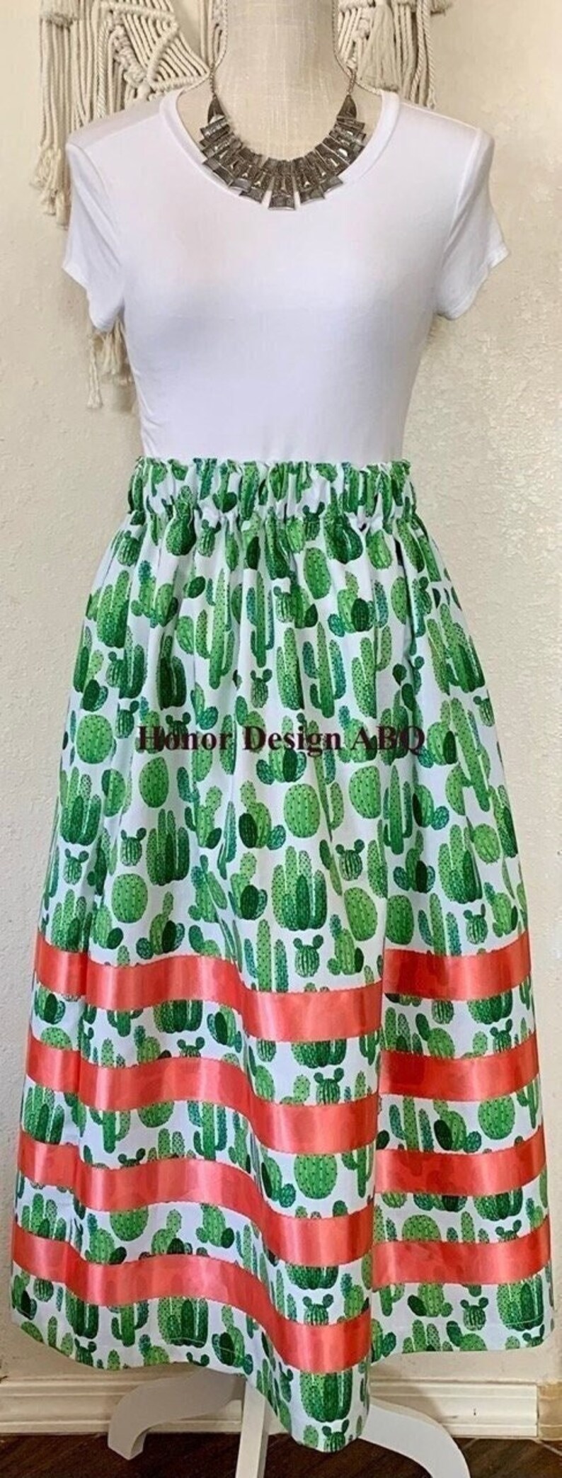 May include: A white skirt with a green cactus print and a coral ribbon trim. The skirt is gathered at the waist and has a flowy silhouette.
