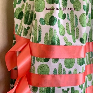 May include: A white fabric skirt with a green cactus print and coral colored ribbon trim. The skirt has the text "Honor Design ABQ" printed on the fabric.