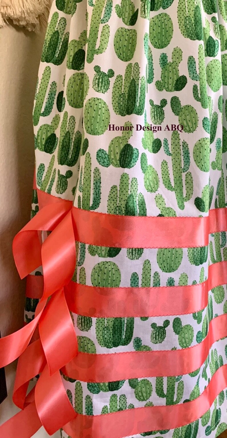 May include: A white fabric skirt with a green cactus print and coral colored ribbon trim. The skirt has the text "Honor Design ABQ" printed on the fabric.