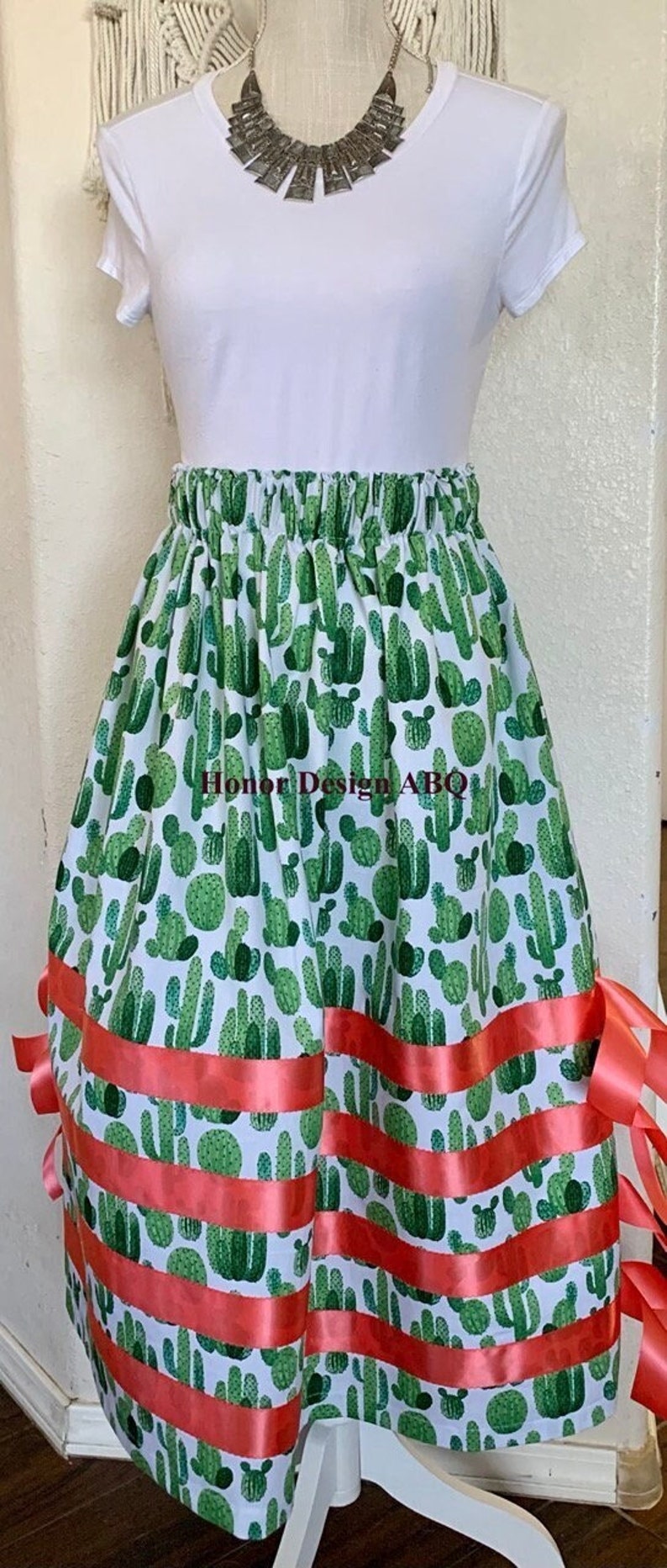 May include: A white skirt with a green cactus print and coral ribbon trim. The skirt has an elastic waistband and is a midi length.