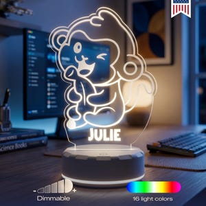 May include: A white LED night light featuring a clear acrylic panel with an etched cartoon monkey holding a banana and the name JULIE printed below. The circular base includes a dimmable light feature with 16 color options and a Made in USA logo.