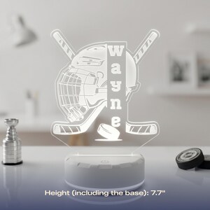 May include: A white hockey-themed LED lamp with the name "Wayne" vertically displayed. The lamp features a hockey helmet, sticks, and a puck design. The base is white, and the height, including the base, is 7.7 inches.