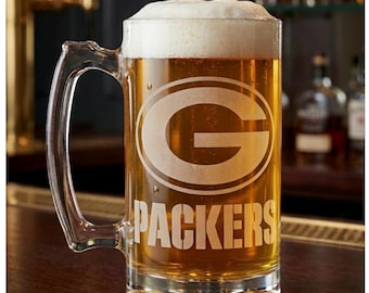 May include: A clear glass beer mug with a sturdy handle, filled with golden amber beer topped with a thick white foam head. The front of the mug features an etched Green Bay Packers logo and the word PACKERS in bold, capital letters.