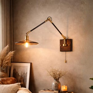 Może przedstawiać: Regulowana lampa ścienna z ciemnobrązowym ramieniem i kloszem oraz akcentami w kolorze mosiądzu. Lampa jest zamontowana na ciemnej drewnianej podstawie. Lampa jest włączona, emitując ciepłą poświatę. Lampa jest częścią sceny wystroju wnętrz.