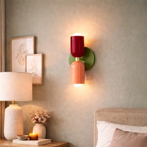 May include: A contemporary wall sconce featuring a red top and a peach-coloured bottom, set on a green circular base. The sconce is lit, emitting a warm light. A bedside table with a lamp, framed art, and a candle is also visible.