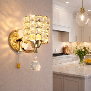 May include: A gold-toned wall sconce with a cylindrical shade composed of square-cut crystals. A crystal ball hangs below the shade. The sconce is mounted on a gold-colored circular base. A small chain with a yellow teardrop-shaped pull hangs from the sconce.