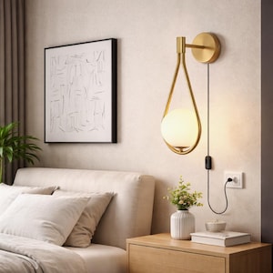 Könnte beinhalten: Goldfarbene Wandleuchte mit tropfenförmigem Design und einer kugelförmigen, mattierten Glaskugel. Die Leuchte ist an einer beigefarbenen Wand montiert, mit einem schwarzen Kabel und Stecker. Ein gerahmtes abstraktes Kunstwerk hängt in der Nähe.