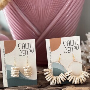 May include: Two pairs of white ceramic earrings with gold hoops. The earrings are displayed on a card with the text "Salty Sea Co." and a website address.