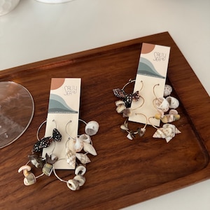 May include: Two pairs of hoop earrings adorned with seashells and small beads. The earrings are displayed on a wooden tray with a white background. Each pair is attached to a card with the text "Salty Sea Co."