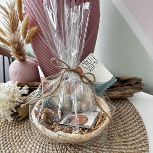 May include: A small wicker basket filled with a variety of items, including a wooden keychain, a pair of earrings, and a small wooden disc with the text "Salty Sea Co.". The basket is wrapped in clear plastic and tied with twine. The basket is sitting on a woven placemat.