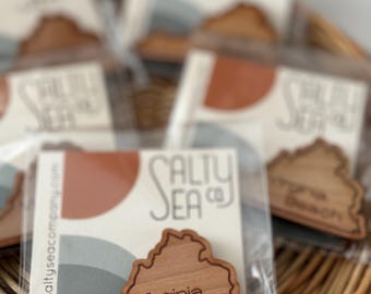 May include: Wooden Virginia Beach souvenir magnets in clear packaging, displayed in a woven basket. Each magnet is shaped like the state of Virginia, with "Virginia Beach" engraved. The packaging features the "Salty Sea Co" logo and website.