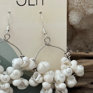 May include: Silver hoop earrings adorned with clusters of small, white seashells. The earrings have a delicate, beachy aesthetic, perfect for adding a touch of coastal charm to any outfit. The earrings are displayed on a neutral background.