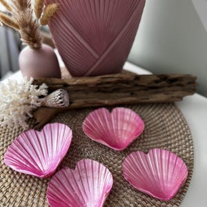 May include: Four pink heart-shaped seashells on a woven mat. The shells are smooth and have a glossy finish.