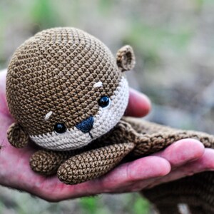 May include: A crocheted otter plush toy in shades of brown and beige. The otter has black eyes and a dark nose. The toy is held in a hand, with the background blurred, suggesting an outdoor setting. The plush is soft and detailed.