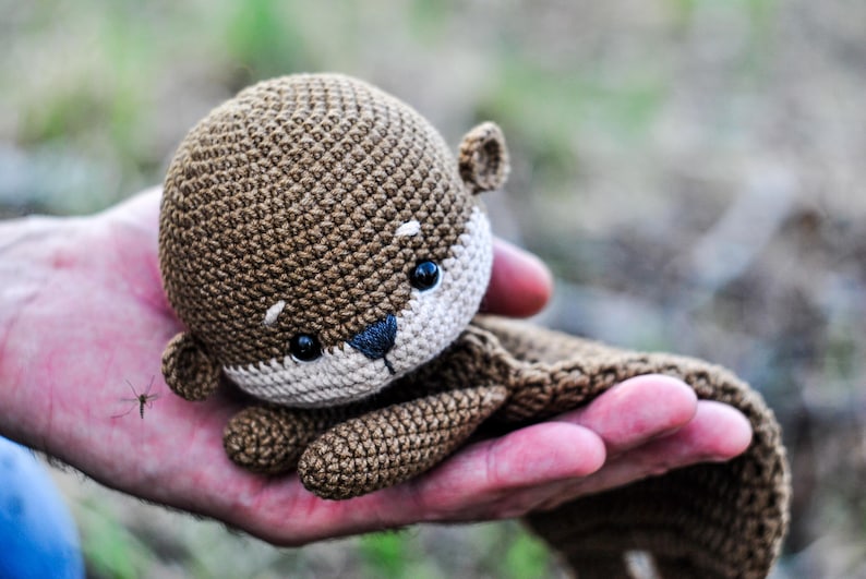 May include: A crocheted otter plush toy in shades of brown and beige. The otter has black eyes and a dark nose. The toy is held in a hand, with the background blurred, suggesting an outdoor setting. The plush is soft and detailed.