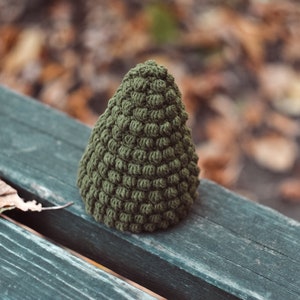 May include: A green crocheted Christmas tree ornament, sitting on a wooden bench with fallen leaves in the background.