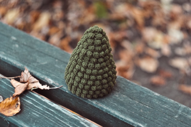 May include: A green crocheted Christmas tree ornament, sitting on a wooden bench with fallen leaves in the background.