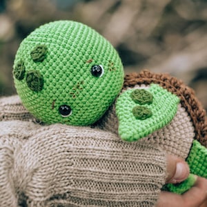 Pu&ograve; includere: Una tartaruga all'uncinetto fatta a mano nei toni del verde, marrone e beige. La tartaruga ha una testa rotonda verde con occhi neri e cuciture decorative. Il guscio &egrave; marrone e il corpo beige. La tartaruga &egrave; tenuta in mano.