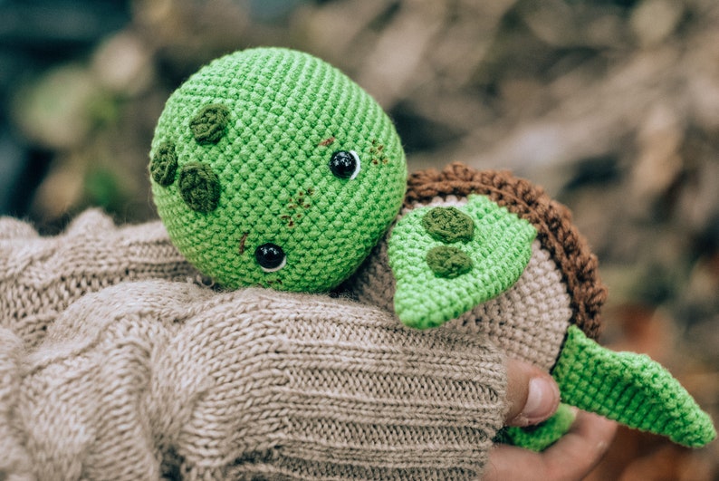 Pu&ograve; includere: Una tartaruga all'uncinetto fatta a mano nei toni del verde, marrone e beige. La tartaruga ha una testa rotonda verde con occhi neri e cuciture decorative. Il guscio &egrave; marrone e il corpo beige. La tartaruga &egrave; tenuta in mano.
