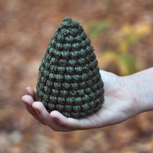 May include: A hand holding a small, dark green crocheted pine tree ornament.