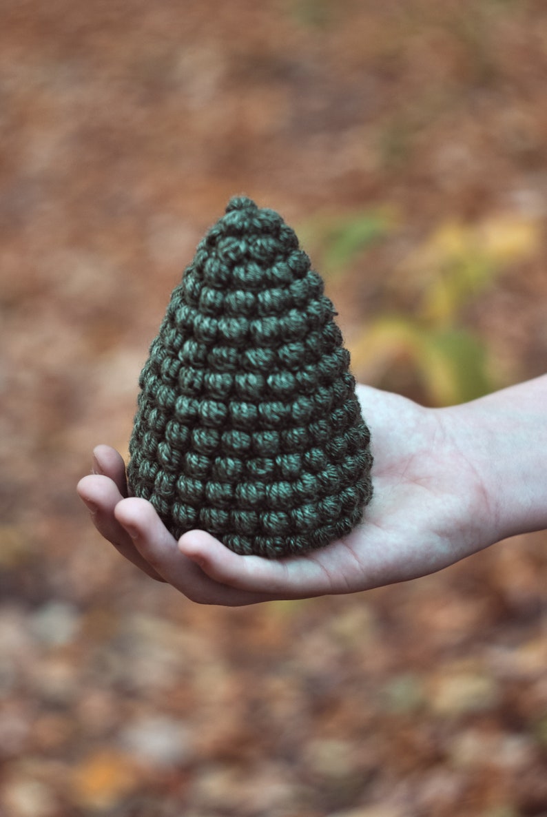 May include: A hand holding a small, dark green crocheted pine tree ornament.