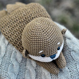 May include: A handmade crochet otter in shades of brown, with a white muzzle and blue nose. The otter is lying on a gray cable-knit blanket. The toy has small black eyes and is made with a tight stitch.