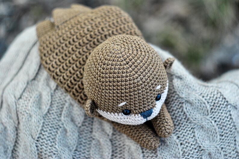 May include: A handmade crochet otter in shades of brown, with a white muzzle and blue nose. The otter is lying on a gray cable-knit blanket. The toy has small black eyes and is made with a tight stitch.