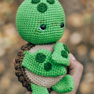 Pu&ograve; includere: Un peluche a forma di tartaruga fatto a mano all'uncinetto. La tartaruga ha testa e arti verdi, guscio marrone e beige e occhi neri. Il peluche &egrave; tenuto in mano, con uno sfondo sfocato di foglie autunnali.