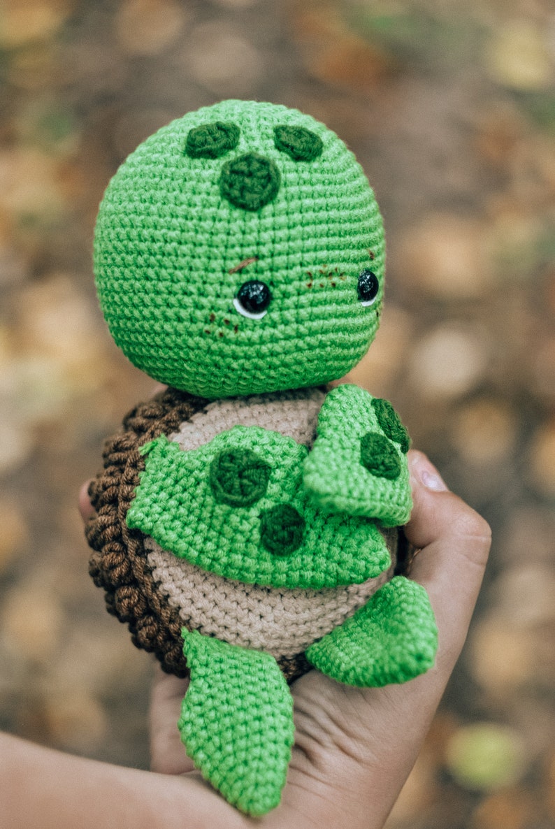 Pu&ograve; includere: Un peluche a forma di tartaruga fatto a mano all'uncinetto. La tartaruga ha testa e arti verdi, guscio marrone e beige e occhi neri. Il peluche &egrave; tenuto in mano, con uno sfondo sfocato di foglie autunnali.