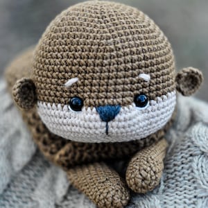 May include: A handmade crochet otter plush toy in shades of brown, beige, and blue. The otter has a rounded head, small ears, and a white face with black eyes and a blue nose. The plush is lying on a gray knitted surface.