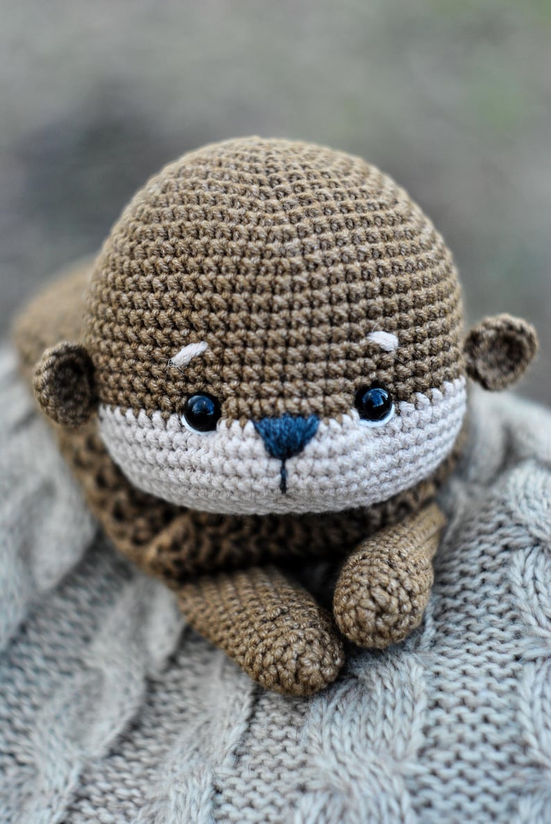 May include: A handmade crochet otter plush toy in shades of brown, beige, and blue. The otter has a rounded head, small ears, and a white face with black eyes and a blue nose. The plush is lying on a gray knitted surface.