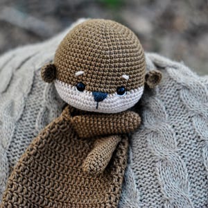 May include: A handmade crochet otter plush toy in shades of brown, beige, and black. The otter has a detailed face with small ears and a long body. It rests on a gray cable-knit blanket, creating a cozy and soft aesthetic.