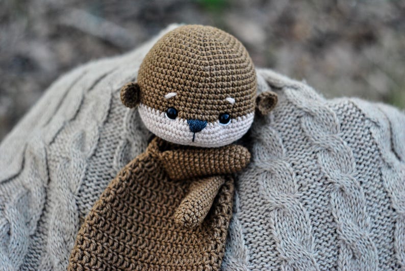May include: A handmade crochet otter plush toy in shades of brown, beige, and black. The otter has a detailed face with small ears and a long body. It rests on a gray cable-knit blanket, creating a cozy and soft aesthetic.