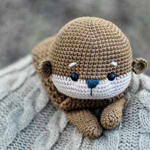 May include: A handmade crochet otter plush toy in shades of brown, beige, and blue. The otter has a detailed, textured design with small ears, a white face, and black eyes. It is lying on a gray, cable-knit blanket.