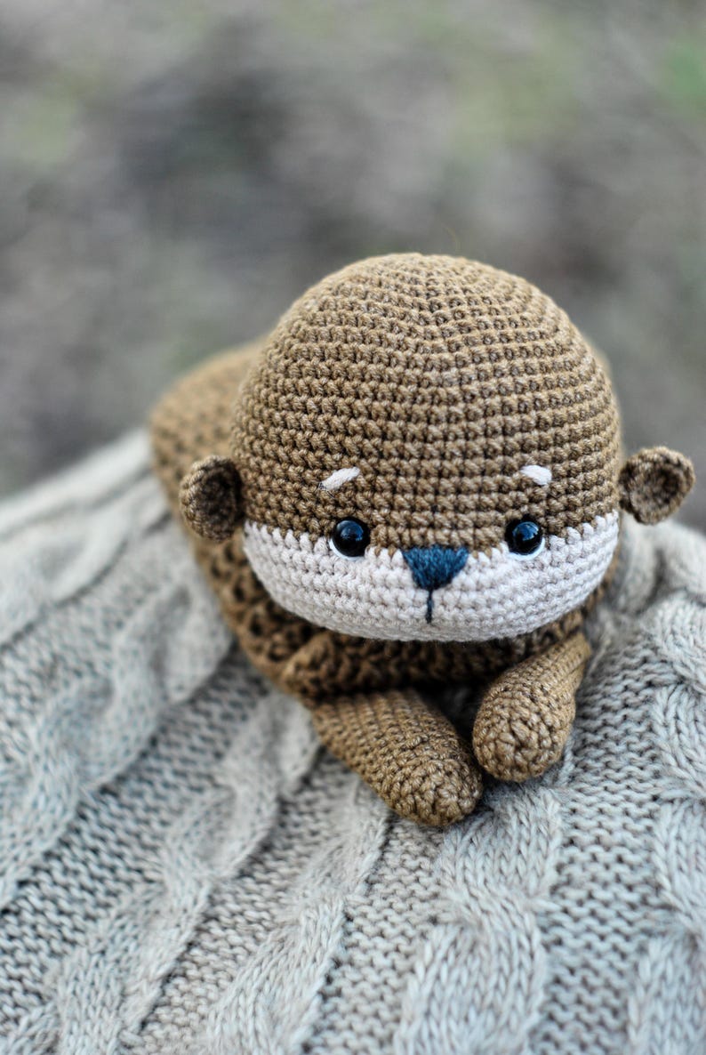 May include: A handmade crochet otter plush toy in shades of brown, beige, and blue. The otter has a detailed, textured design with small ears, a white face, and black eyes. It is lying on a gray, cable-knit blanket.