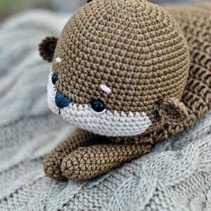 May include: A handmade crochet otter plush toy in shades of brown and beige. The otter has a detailed, textured body, small ears, and a white muzzle with a blue nose. The toy is lying on a gray, cable-knit blanket.
