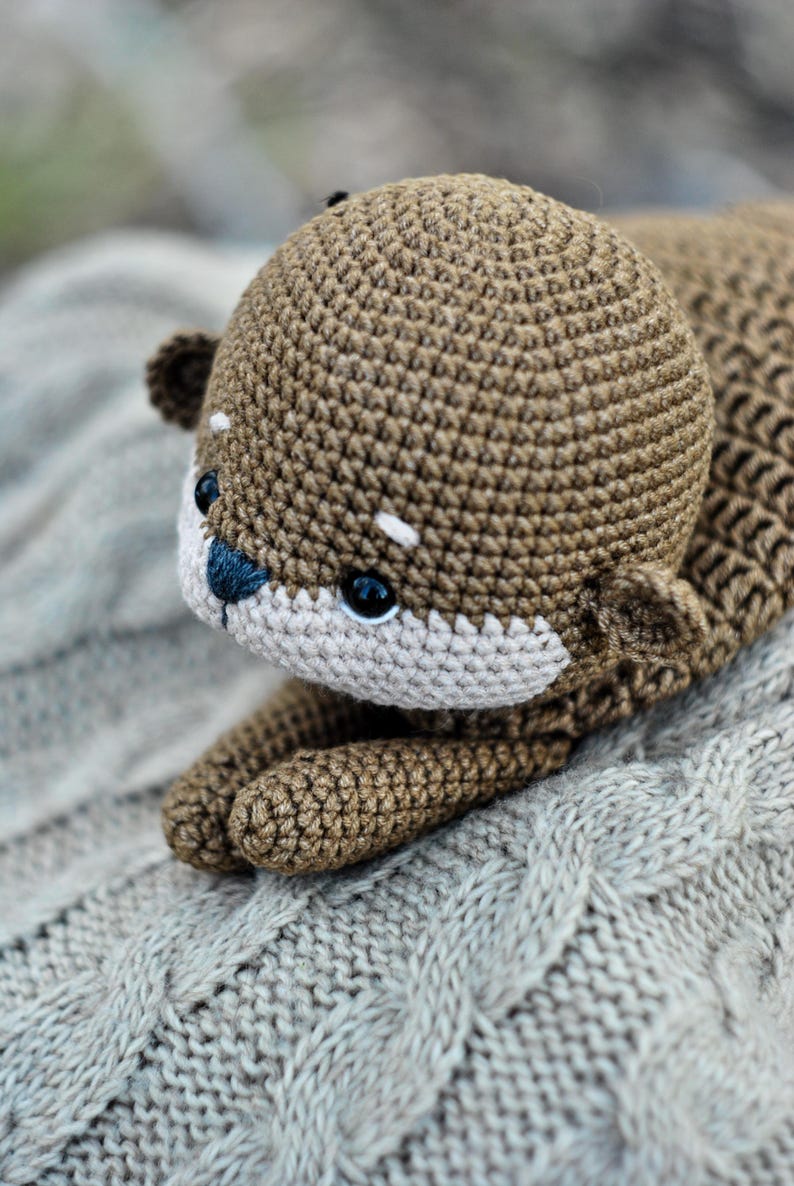 May include: A handmade crochet otter plush toy in shades of brown and beige. The otter has a detailed, textured body, small ears, and a white muzzle with a blue nose. The toy is lying on a gray, cable-knit blanket.