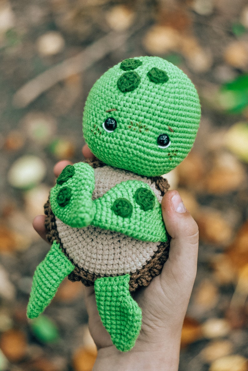 Pu&ograve; includere: Una tartaruga verde all'uncinetto fatta a mano con un guscio marrone chiaro e finiture marrone scuro. La tartaruga ha occhi neri e macchie verdi sulla testa e sulle pinne. La tartaruga &egrave; tenuta nella mano di una persona.