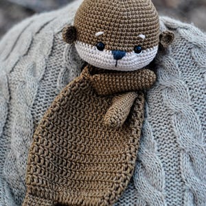 May include: A handmade crochet otter plush toy in shades of brown and cream. The otter has a detailed, textured design with small ears, a cream-colored face, and black eyes. The plush is lying on a gray cable-knit blanket.